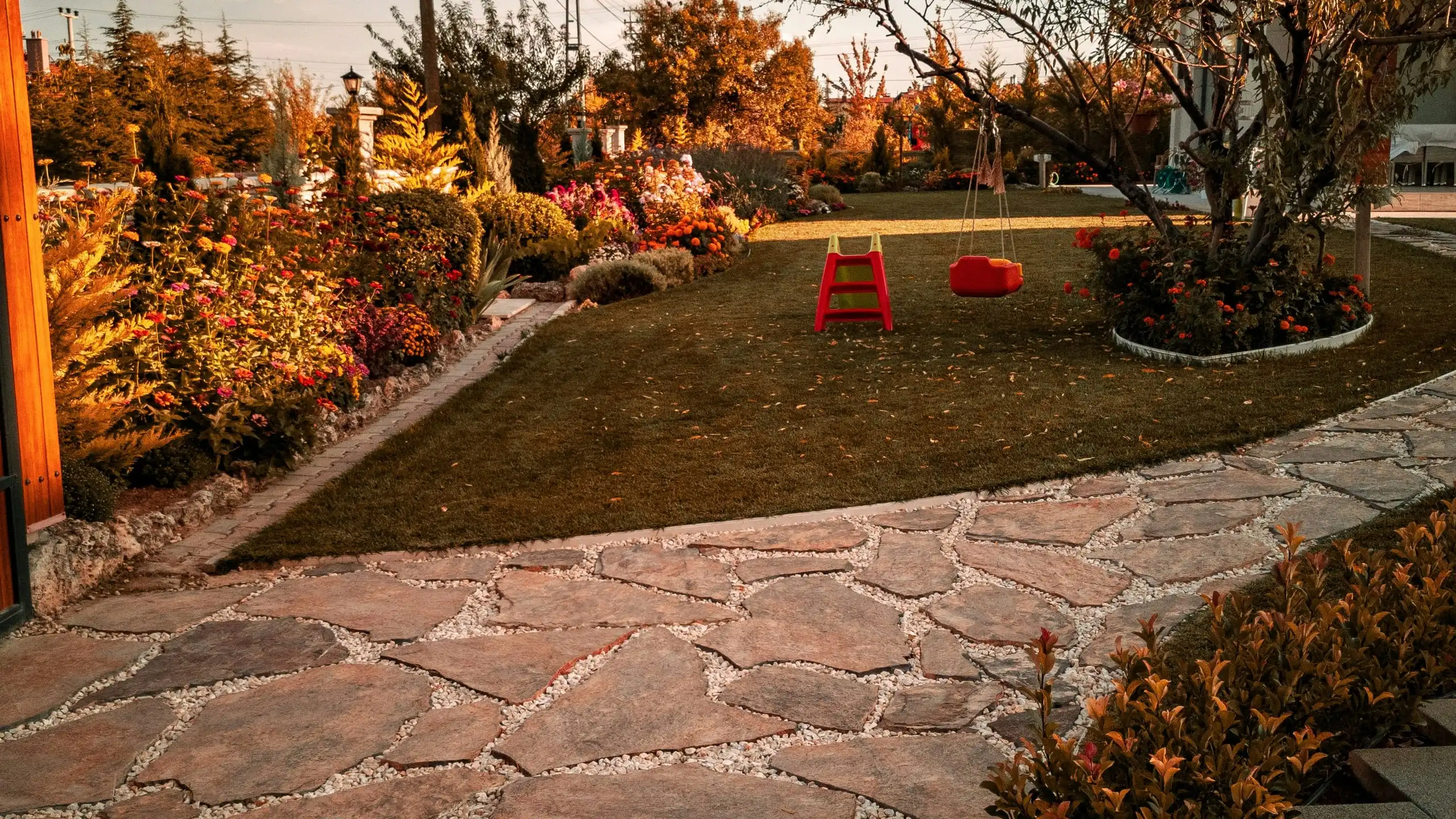 A flagstone path beside a lawn with two red swings hung from a tree, garden beds along the fence